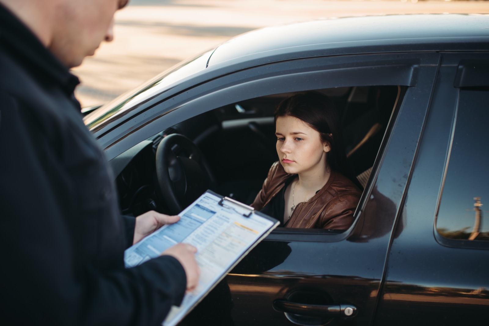 An officer stands at the driver's window writing on a clipboard while the driver waits, hands in view.